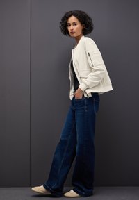 Vit ribbad bomberjacka, svart topp, oversized mörkblå denimjeans, beige skor. Modellen står mot en mörkgrå bakgrund.