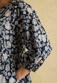 Eine Nahaufnahme eines dunkelblauen Kleides einer Frau mit weißen Blumendrucken. Ihre Hand ruht in der Tasche und zeigt den elastischen Puffärmel des Kleides.