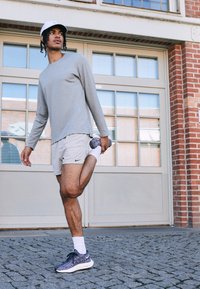 Model Nike Sportswear: šedé tričko s dlouhým rukávem, béžové šortky, vzorované černobílé tenisky, natahující jednu nohu, městské pozadí, dlážděná podlaha.