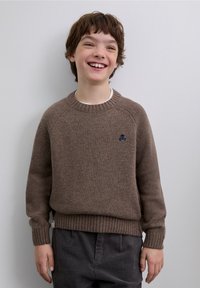Brown knitted sweater with ribbed cuffs and hem, featuring a small blue skull emblem on the chest. Worn with gray corduroy pants.