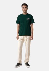 Dark green cotton t-shirt with short sleeves and a front graphic design, paired with off-white pants and black sneakers.
