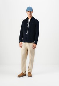 Cămașă button-up de culoare albastru închis, pantaloni bej și pantofi maro. Căciulă de culoare albastru deschis. Design minimalistic, material Neted și branding subtil.