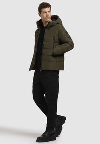Jachetă puffer de culoare verde măslin cu glugă, având buzunare cu fermoar și un design matlasat. Pantaloni cargo negri și cizme completează ținuta.