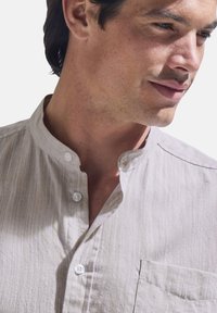 Light beige, collared shirt with a buttoned placket, vertical stripes, and a chest pocket. Fabric appears soft with a textured finish.