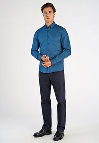 Blauwe button-up shirt met een gestructureerde afwerking, zwarte knopen en lange mouwen, gecombineerd met donkerblauwe broek en zwarte schoenen.