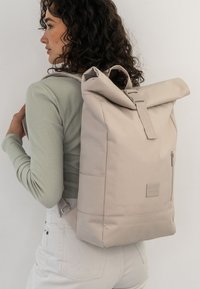 Beige hengeres hátizsák, textúrált anyagból, oldalsó cipzáros zsebbel és állítható pántokkal, minimalista dizájnjával és puha éleivel.