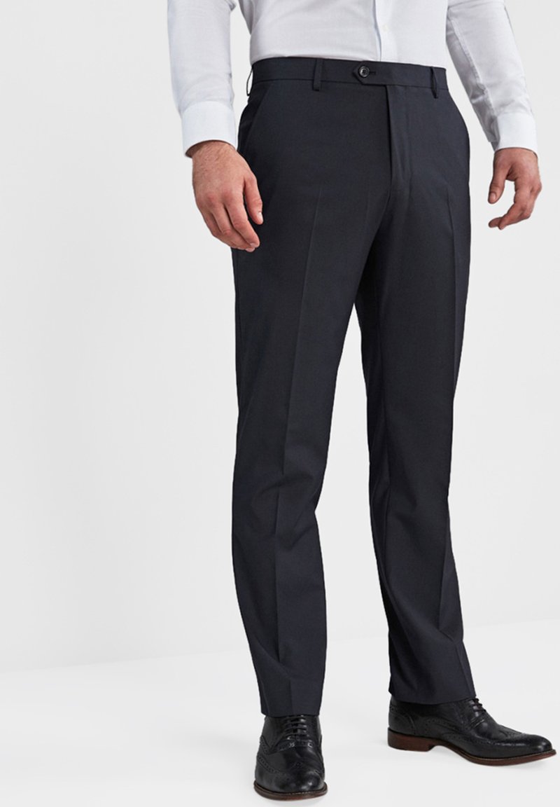 Op maat gemaakte donkerblauwe broek in een gladde stof met een platte voorkant, riemlussen en een enkele knoopsluiting, gecombineerd met zwarte dresschoenen.