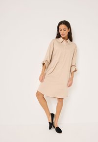 Beige gombos ruha gallérral és felgombolt ujjal, lazább szabással és sima textúrával, fekete slip-on cipővel párosítva.