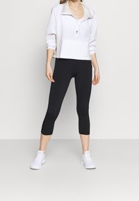 Veste blanche à fermeture éclair avec une poche avant, associée à des leggins capri noirs et des baskets blanches, posant devant un fond neutre.