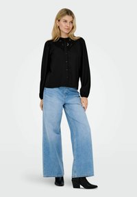 Donna che indossa una blusa nera a maniche lunghe con dettagli in pizzo, jeans larghi azzurri e stivaletti neri con tacco, in piedi su uno sfondo bianco.