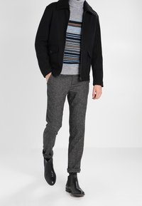 Černá bunda s kožíškovým límcem, šedý proužkovaný svetr, šedé texturované kalhoty a černé kotníkové boty, zvýrazňující vrstvení látek a barevný kontrast.