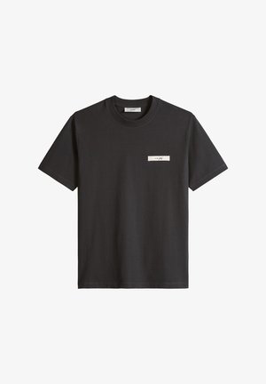 Czarny T-shirt z krótkim rękawem i okrągłym dekoltem, z małą białą prostokątną naszywką z kursywnym tekstem na lewej piersi.