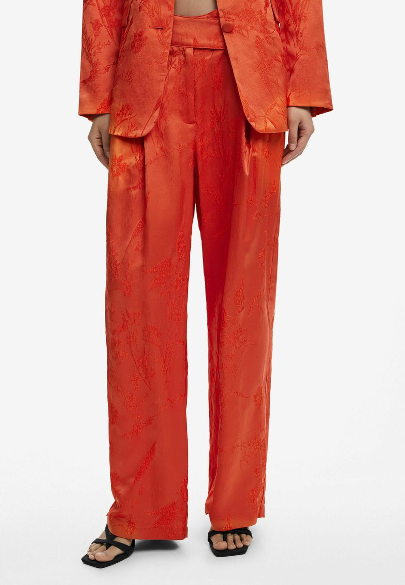 Traje de satén rojo con un patrón floral texturizado. Los pantalones de pierna ancha tienen una cintura alta y un corte relajado, complementados con sandalias negras con tiras.