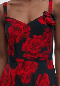 Κοντινό πλάνο μαύρου φορέματος με μεγάλα, ζωηρά κόκκινα τυπώματα τριαντάφυλλων, με αμάνικη σχεδίαση και λεπτομέρεια από ύφασμα τριαντάφυλλο στον αριστερό ιμάντα.