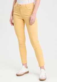 Jean skinny jaune avec une texture lisse, cinq poches et une fermeture à bouton, associé à des baskets blanches avec une semelle beige.
