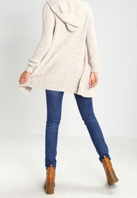 Beige huva cardigan med texturerad stickning, öppen framsida och långa ärmar, tillsammans med mörkblå denimjeans och bruna klackstövlar.
