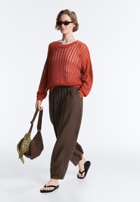 Femme portant un pull en maille orange rouille, pantalon marron ample, sandales noires, lunettes de soleil, tenant un sac marron avec une écharpe verte.