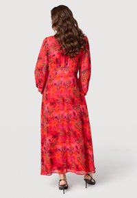 Maxi-jurk in een levendig rood met bloemenpatronen in roze, zwart en groen. Doorzichtige lange mouwen en een getailleerde taille. Draagt zwarte hakken.