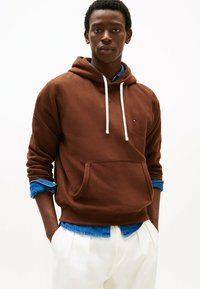 Felpa con cappuccio marrone in tessuto morbido, con cordini bianchi, tasca frontale e polsini a costine, indossata sopra una camicia di jeans blu.