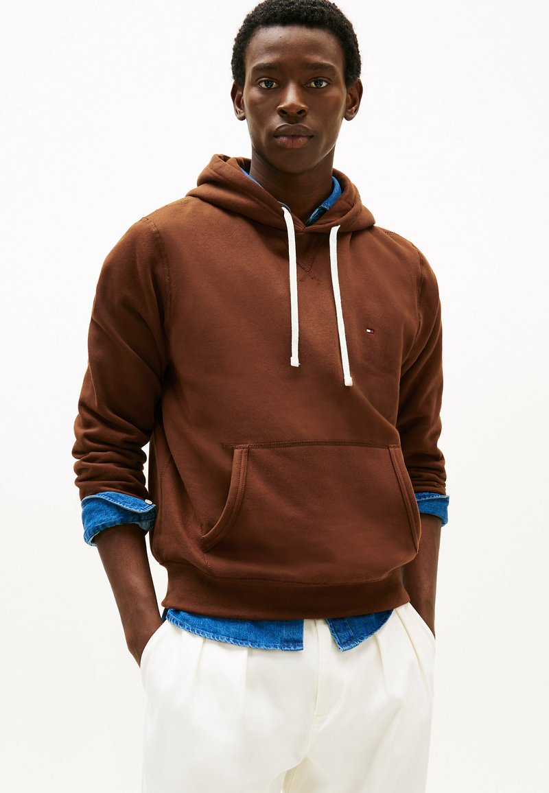 Felpa con cappuccio marrone in tessuto morbido, con cordini bianchi, tasca frontale e polsini a costine, indossata sopra una camicia di jeans blu.