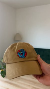 Világosbarna baseball sapka, az elején kerek, színes hímzéssel, amely kék kezet és arcot ábrázol. Ívelt karimával és állítható hátsó pánttal rendelkezik.