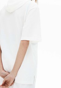 Vit kortärmad topp med luva och texturerat tyg, sidospjäll för rörelse och en avslappnad passform. Bakifrån syns händerna knäppta bakom ryggen.