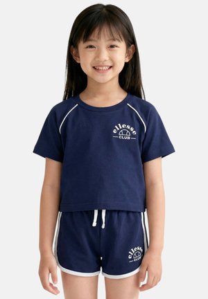 Fille souriante aux longs cheveux foncés porte un short de sport bleu marine et un haut à manches courtes, chacun avec le logo blanc "ellesse CLUB".