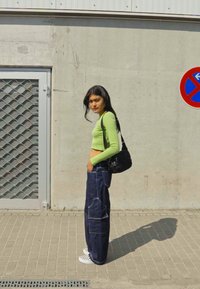 Světle zelený dlouhý rukáv crop top, tmavě modré široké džíny s kapsami, černá ramenní taška, bílé kostkované tenisky, stojící u zdi.