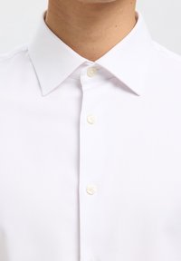 Primer plano de una persona que lleva una camisa blanca de vestir con cuello puntiagudo y tres botones blancos visibles abrochados al frente.
