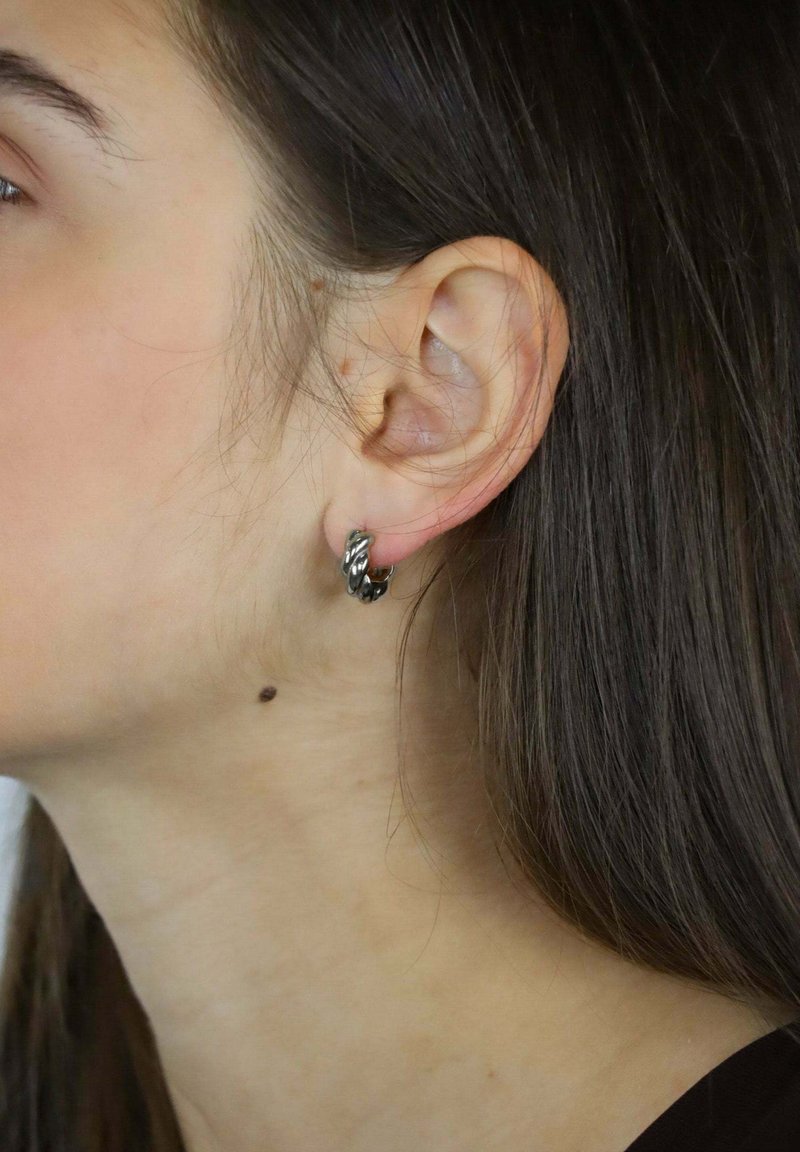 Srebrne kolczyki koła z przekręconym wzorem, z błyszczącą powierzchnią i małym zapięciem zatrzaskowym, noszone na lewym uchu.