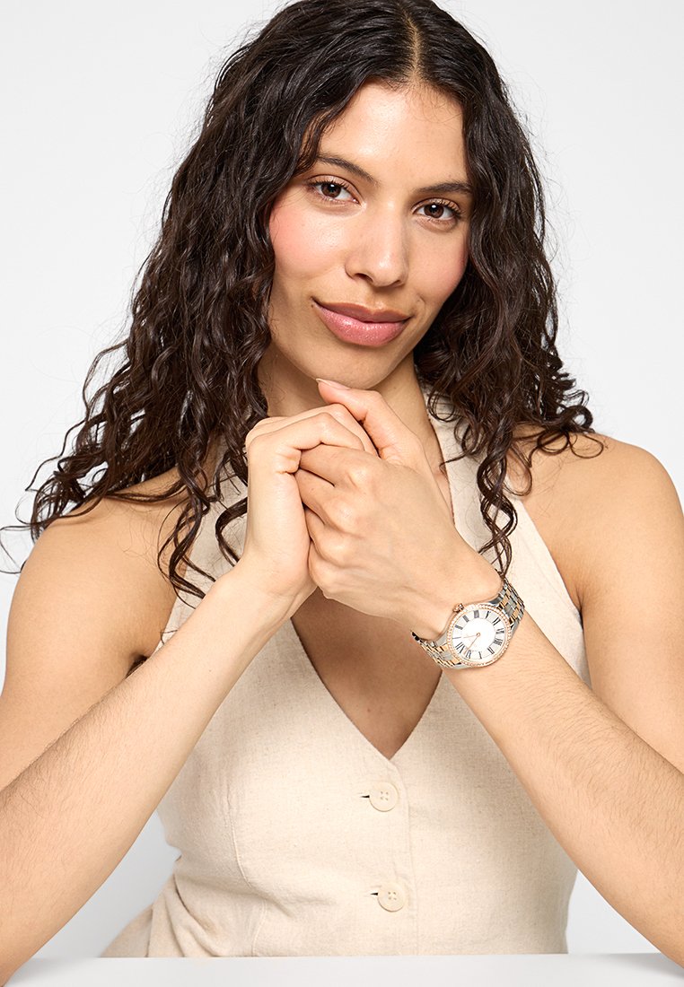 Srebrna metalowa zegarek z białą tarczą, złotymi akcentami i niebieskimi znacznikami godzin, noszony na nadgarstku. Modelka ma długie, kręcone włosy i siedzi.