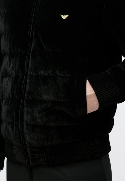 Μαύρο μπουφάν Quilted puffer με απαλό, βελούδινο υφή, διαθέτει ριμπ μανσέτες και μικρό χρυσό λογότυπο στο στήθος. Τσέπη με φερμουάρ στο μπροστινό μέρος.