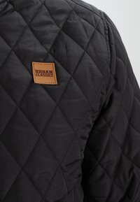 Zwarte gewatteerde jas met diamantpatronen, een zachte textuur en een bruine leren logo-patch met het label "URBAN CLASSICS."