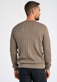 Brun kabelstickad tröja med ribbade ärmslut och nederkant, med ett strukturerat mönster och rundad hals, kombinerad med mörka byxor.