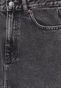 Gros plan sur un jean en denim noir montrant la taille, le bouton en métal, la passoire pour ceinture et la poche avant avec des détails de couture visibles.