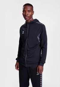 Zwarte rits hoodie met capuchon, mesh textuur en grijze accenten. Bevat het hummel-logo en contrasterende strepen op de mouwen.