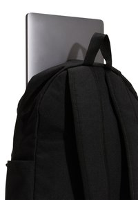 Svart tygryggsäck med vadderade remmar; en tunn, silverfärgad laptop delvis inskjuten i bakfickan. Minimalistisk design och textur.