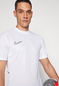 Hvit sportsskjorte med korte ermer, rund hals og svart Nike-logo. Holder en svart-hvit mønstrete fotball med røde detaljer.