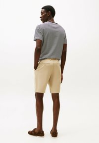 Bărbat îmbrăcat cu un tricou cu dungi alb-negru, pantaloni scurți bej și pantofi casual maro, stând cu mâinile în buzunare, întors cu fața înapoi.