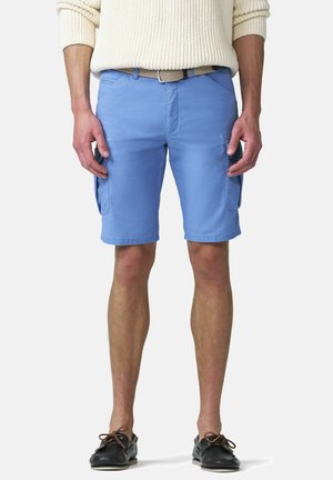 Mann trägt hellblaue Cargoshorts, einen cremefarbenen Strickpullover, einen beigefarbenen geflochtenen Gürtel und schwarze Bootsschuhe, steht vor einem einfarbigen Hintergrund.