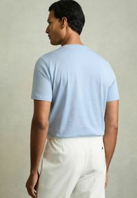 Tricou de culoare albastru deschis, cu mânecă scurtă, textură netedă, guler rotund și croială lejeră, purtat cu pantaloni albi ajustați.