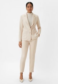 Blazer beige su misura con due tasche frontali e chiusura a bottoni, abbinato a pantaloni slim coordinati. Tessuto liscio e design aderente.