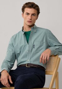 Joven con cabello castaño claro y ojos azules, vistiendo una camisa verde a cuadros, pantalones oscuros y un cinturón trenzado marrón, sentado en una silla de madera.