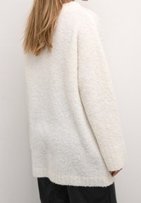 Pull blanc surdimensionné en maille texturée, avec manches longues et ourlet arrondi, porté de manière décontractée avec un bas foncé.