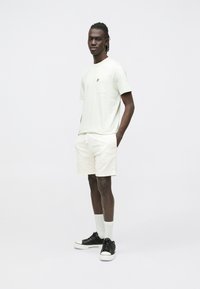 Light green short-sleeve t-shirt with pocket and small logo, paired with textured white shorts and black sneakers with white soles.