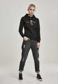 Felpa nera con cappuccio e tasca frontale, presenta una grafica colorata di "Topolino". Indossata con jeans scuri strappati e sneakers alte nere.