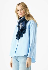 Écharpe bleu marine avec des bords froncés, présentant un détail rayé bleu clair et rose. Portée par-dessus une chemise bouton exprimant une teinte bleu clair.