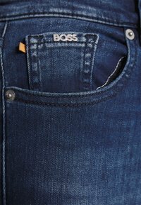 Jeans de denim azul con un pequeño bolsillo frontal que presenta un logo plateado de "BOSS" y costuras en contraste. La tela tiene una textura suave.