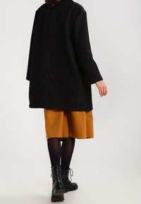 Donna che si allontana indossando un cappotto nero, una gonna giallo senape, collant neri e stivaletti neri con lacci su uno sfondo semplice.