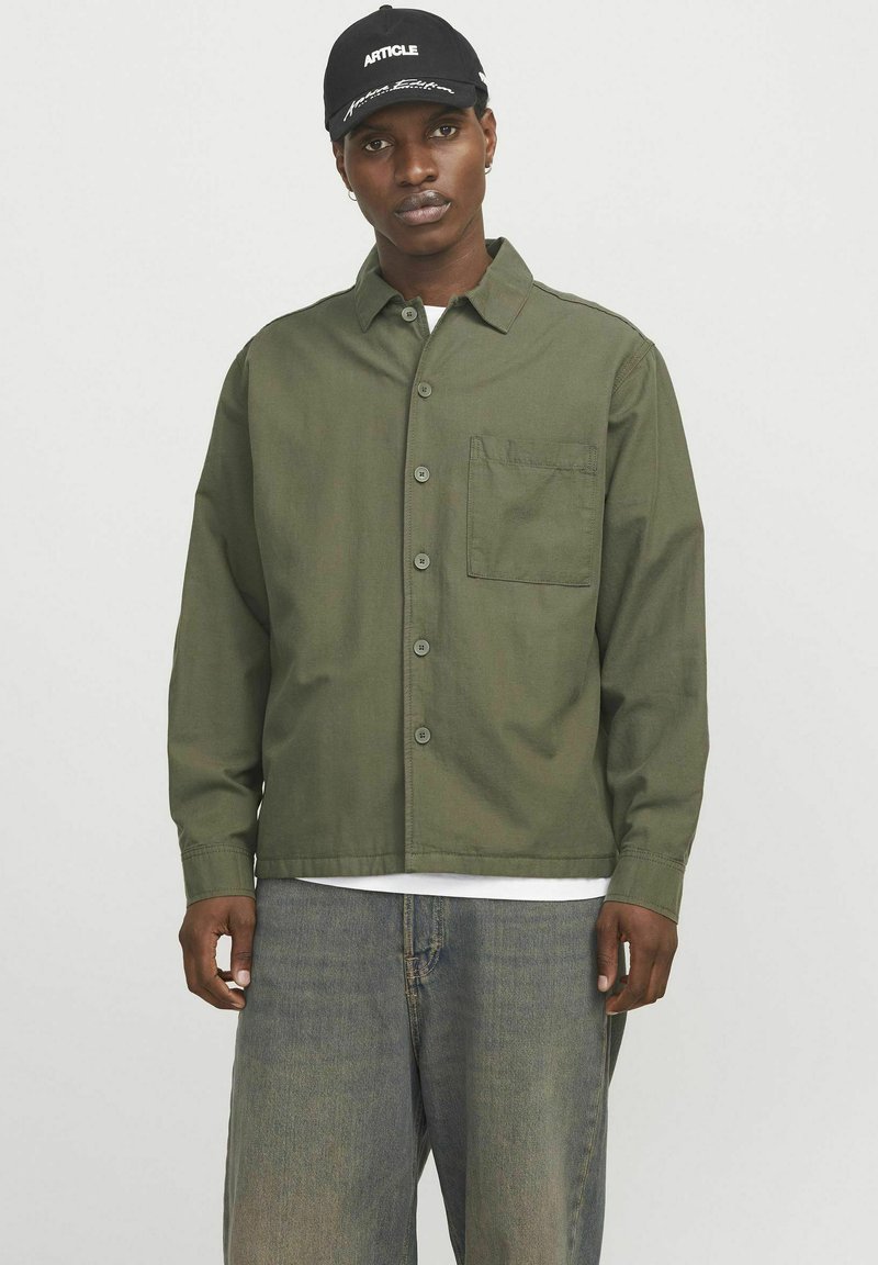 Chemise olive à boutons avec une poche poitrine, en tissu lisse. Associée à un denim bleu clair. Casquette noire avec un texte blanc.